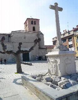 Cruz de piedra en una plaza con árbol podado y edificios en el fondo.