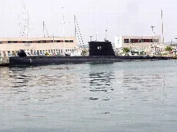 Black submarine in water with buildings and boat masts in the background.