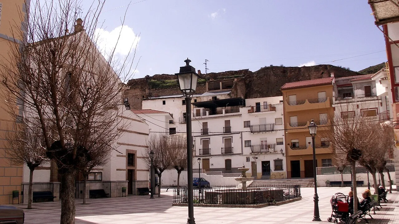 Plaza con árboles, farolas y edificios blancos y beige en Zújar, Granada.