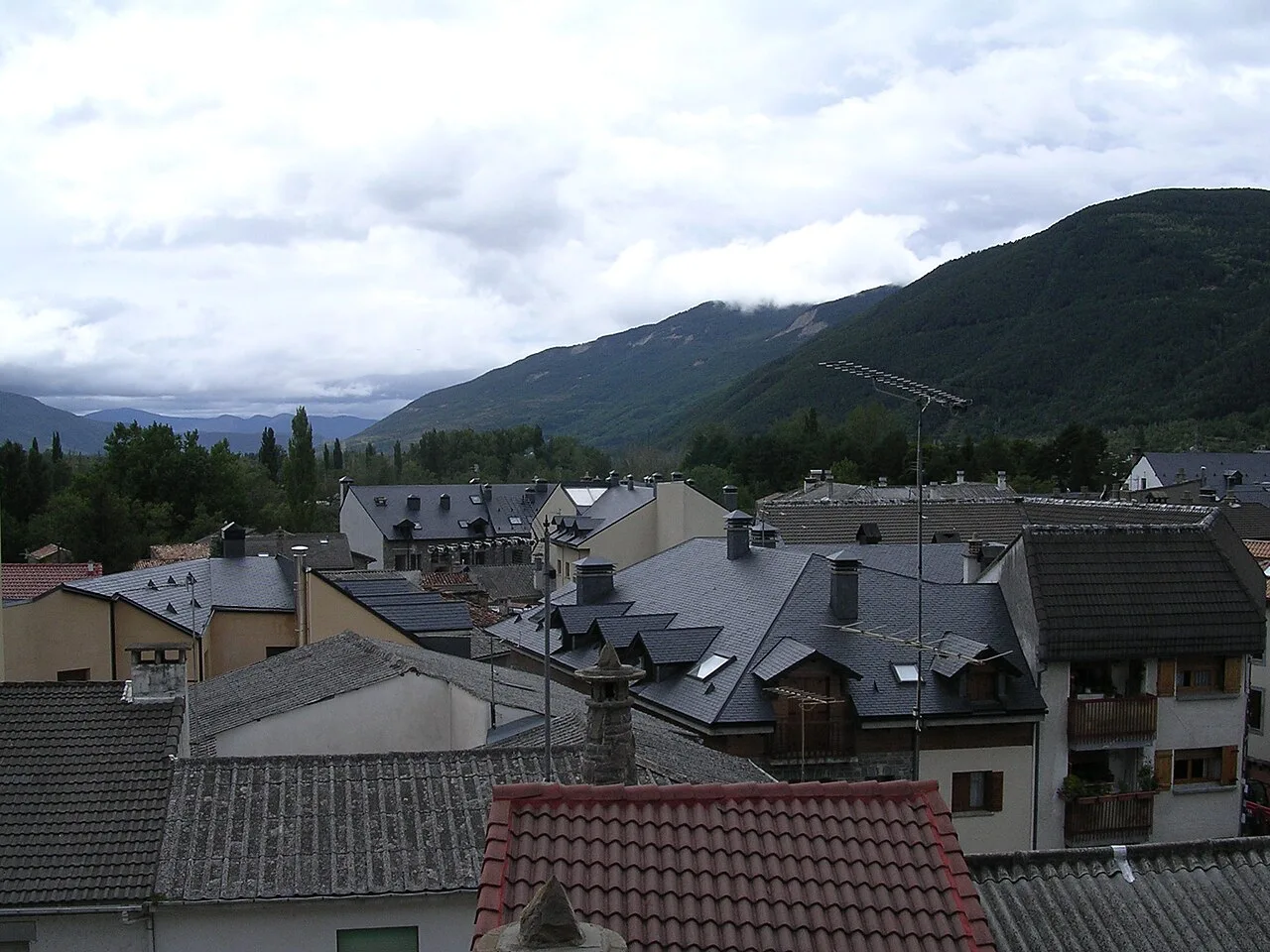 Häuser und Dächer in Biescas mit Bergen und bewölktem Himmel.