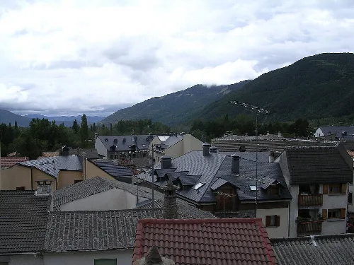 Häuser und Dächer in Biescas mit Bergen und bewölktem Himmel.