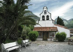 White church with bell tower and red tiled roof, surrounded by greenery and mountains.