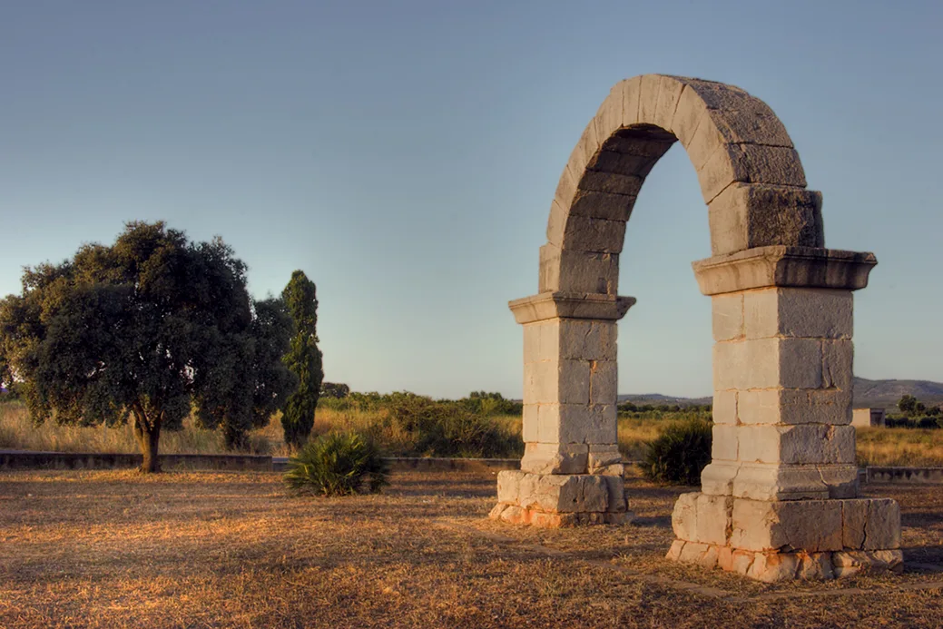 Stone Roman arch in a field with trees and clear sky.