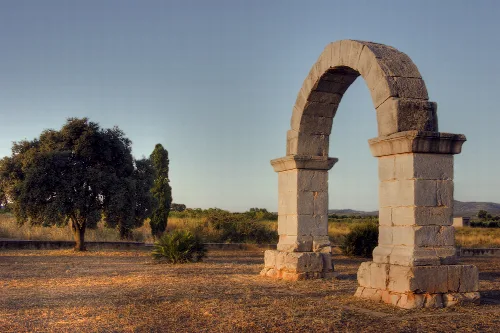 Stone Roman arch in a field with trees and clear sky.