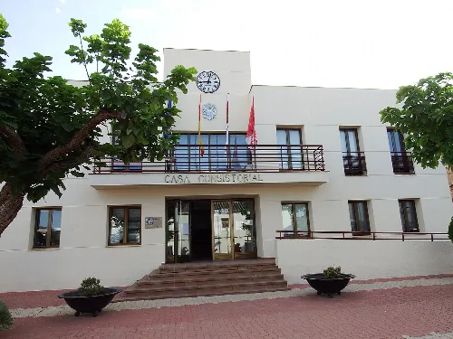 White building with clock and flags, entrance with steps and two flowerpots in front.