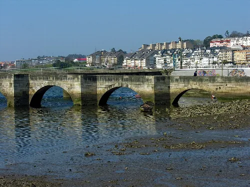 Puente de piedra sobre el río en Cambre con edificios en el fondo.