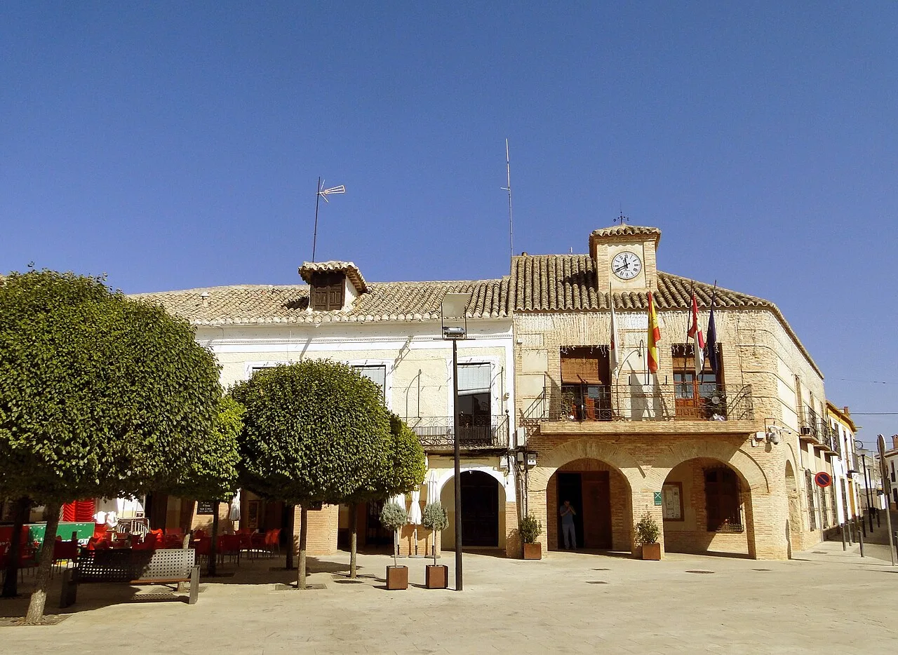 Plaza con árboles, bancos y edificio con reloj y banderas en Carrión de Calatrava.
