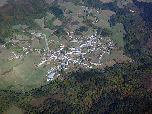 Vista aérea de un pueblo con edificios y caminos en un valle rodeado de árboles.