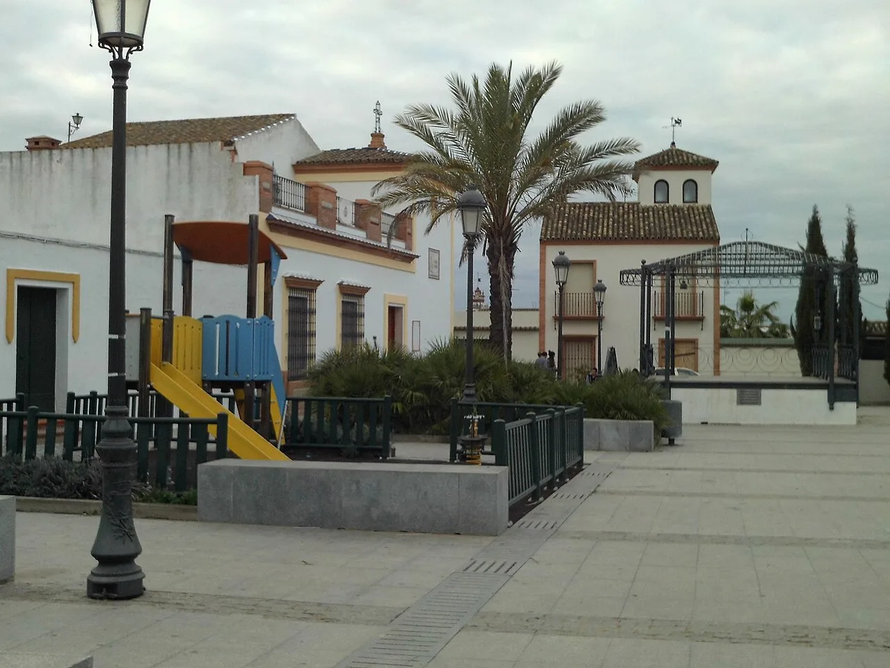 Plaza con juegos infantiles, palmeras y una iglesia en Villanueva del Ariscal.