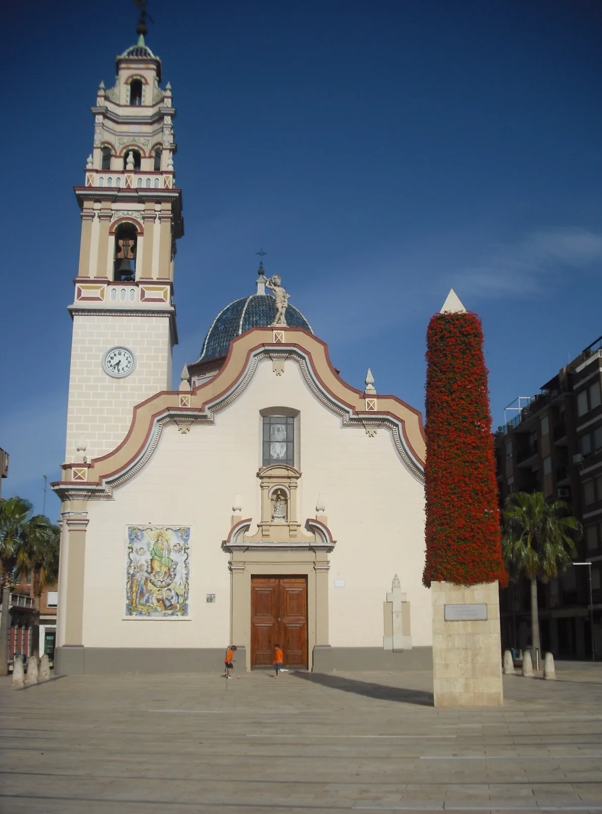 Fachada de la iglesia con un obelisco cubierto de flores rojas a la derecha.