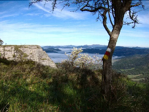 Wanderwegmarkierung an einem Baum in Arrastaria, Álava, mit Bergen und Seen im Hintergrund.