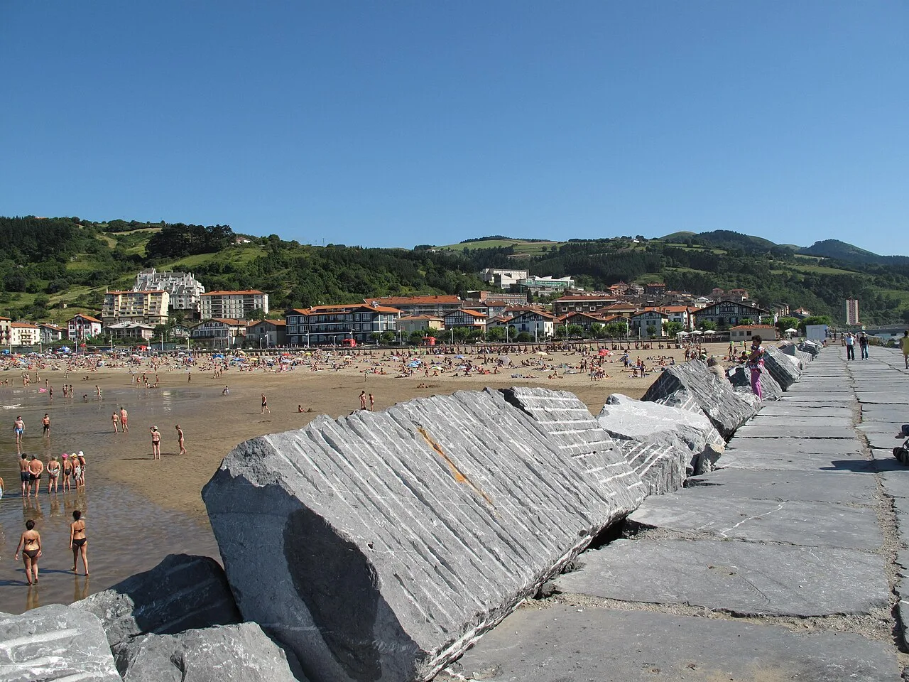 Felsen im Vordergrund und Strand mit Menschen und Gebäuden in Deva, Guipúzcoa.