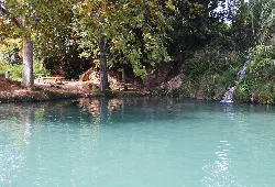 Turquoise waters in a pond surrounded by trees and a small waterfall.