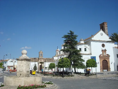 Fuente y iglesia en una plaza con árboles y personas en Palma del Río.