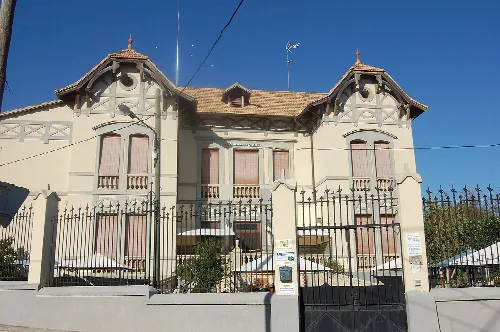 Edificio con fachada blanca y rejas negras, con tejado de tejas y ventanas con cortinas.