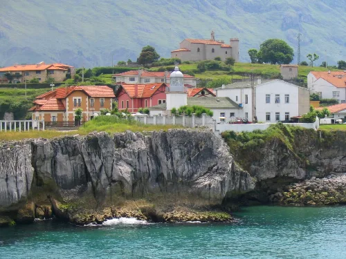 Faro en el puerto de Llanes, casas y castillo en un paisaje costero.