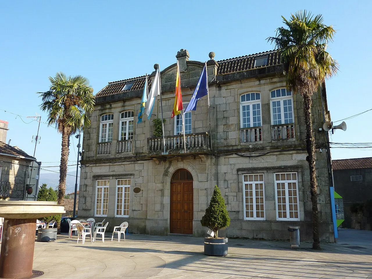 Edificio del ayuntamiento de Arbo con banderas y palmeras.