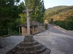 Cruz de piedra en la orilla de un camino con árboles y montañas al fondo.