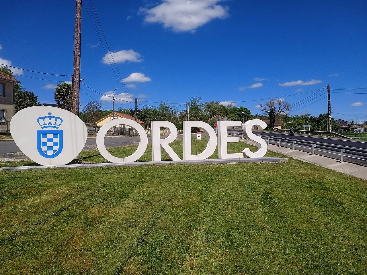 Großes Schild mit dem Namen und Wappen von Órdenes in ländlicher Umgebung.