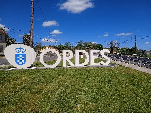 Cartel grande con el nombre y escudo de Órdenes en un entorno rural.