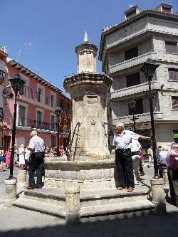 Steinbrunnen im Platz mit Menschen und Gebäuden drumherum.