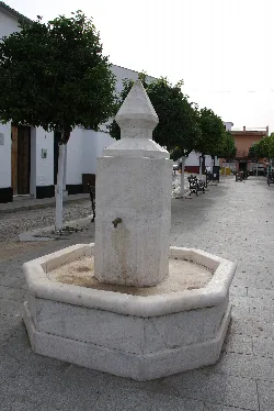 Fuente de piedra con forma de columna en una plaza con árboles y bancos.