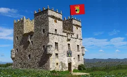 Stone castle with flag and coat of arms on top, in a rural landscape with blue sky.
