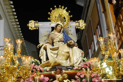 Escultura de la Virgen de las Angustias con Jesús en brazos, rodeada de candelabros dorados y flores rosas.