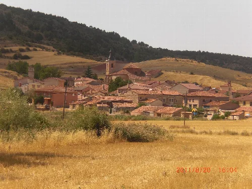 Panoramic view of a village with houses and a church in the center, surrounded by hills and fields.