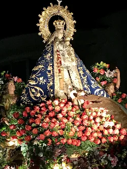 Bild der Virgen de Riánsares mit Krone und blauer Mantel, umgeben von Rosen und anderen Blumen.