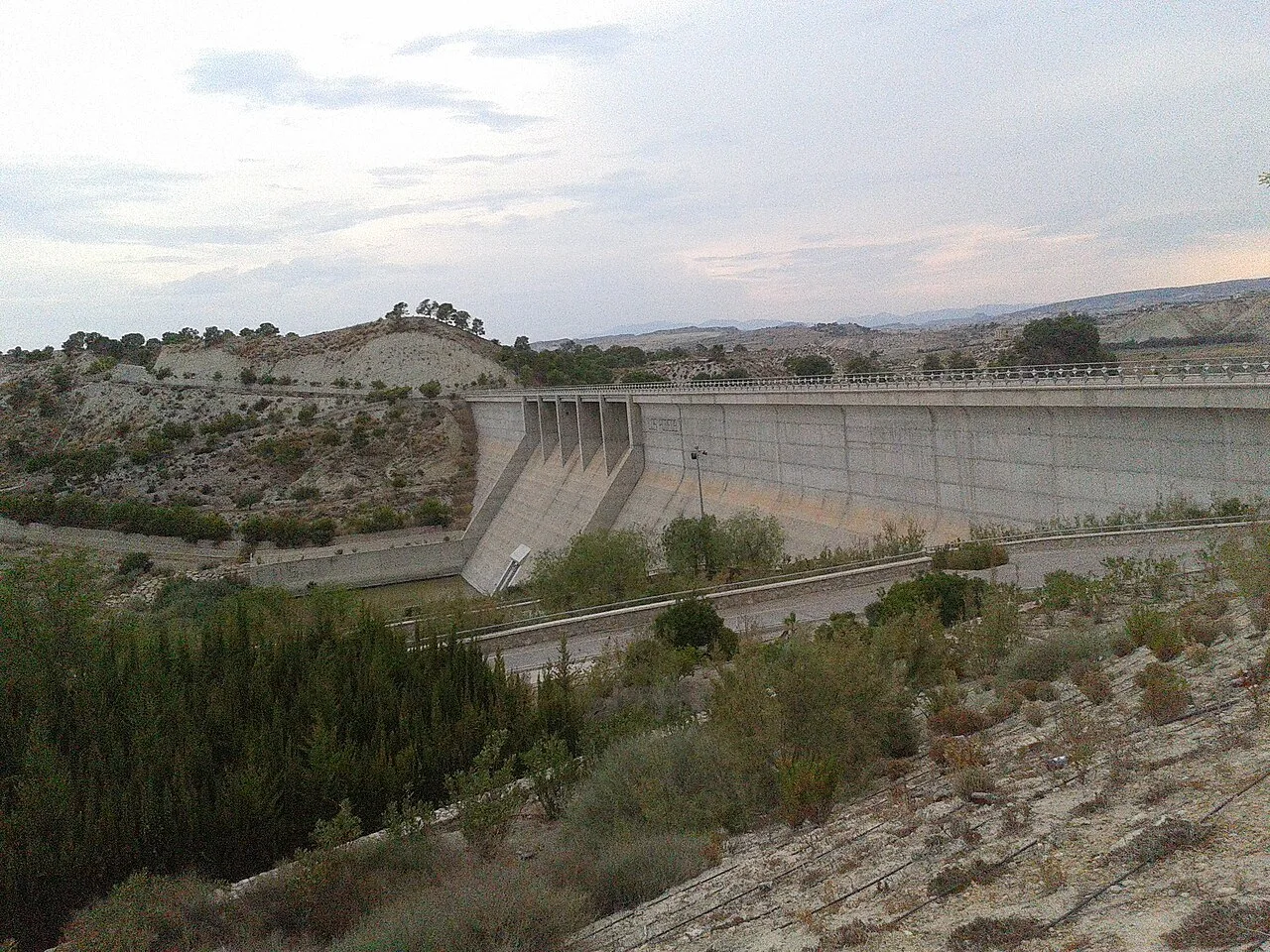 Large dam in a dry landscape with vegetation and hills.