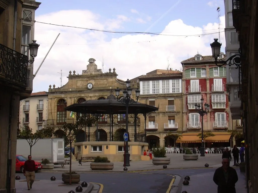 Plaza con edificios antiguos, quiosco en el centro y personas paseando.
