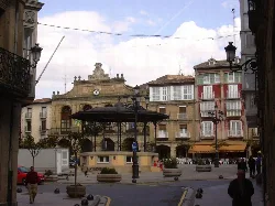 Plaza con edificios antiguos, quiosco en el centro y personas paseando.