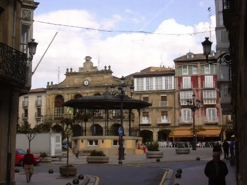 Plaza con edificios antiguos, quiosco en el centro y personas paseando.