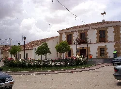 Square with trees, flowers, and a two-story building in Vadillo de la Guareña.