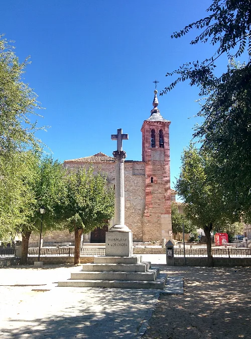 Steinkreuz in einem Platz mit Bäumen und Kirche im Hintergrund.