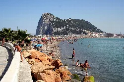 Playa con arena, rocas y gente en el agua, vista del Peñón de Gibraltar al fondo.