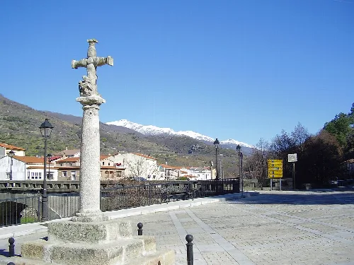 Steinkreuz in einem Platz von Navaconcejo mit schneebedeckten Bergen und blauem Himmel.