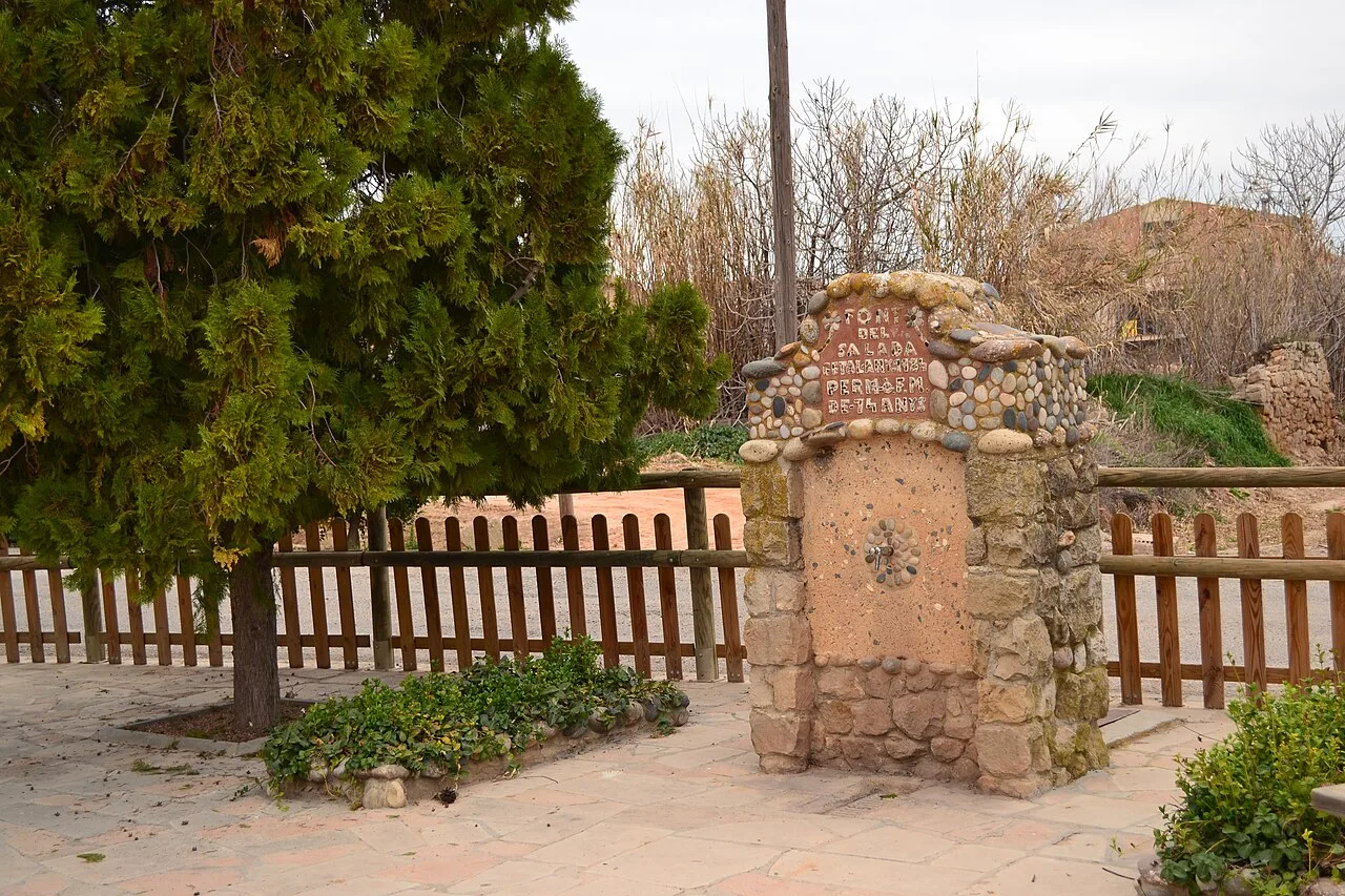 Fuente de piedra con inscripción en Alcanó, rodeada de árboles y plantas.