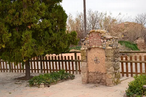Steinbrunnen mit Inschrift in Alcanó, umgeben von Bäumen und Pflanzen.