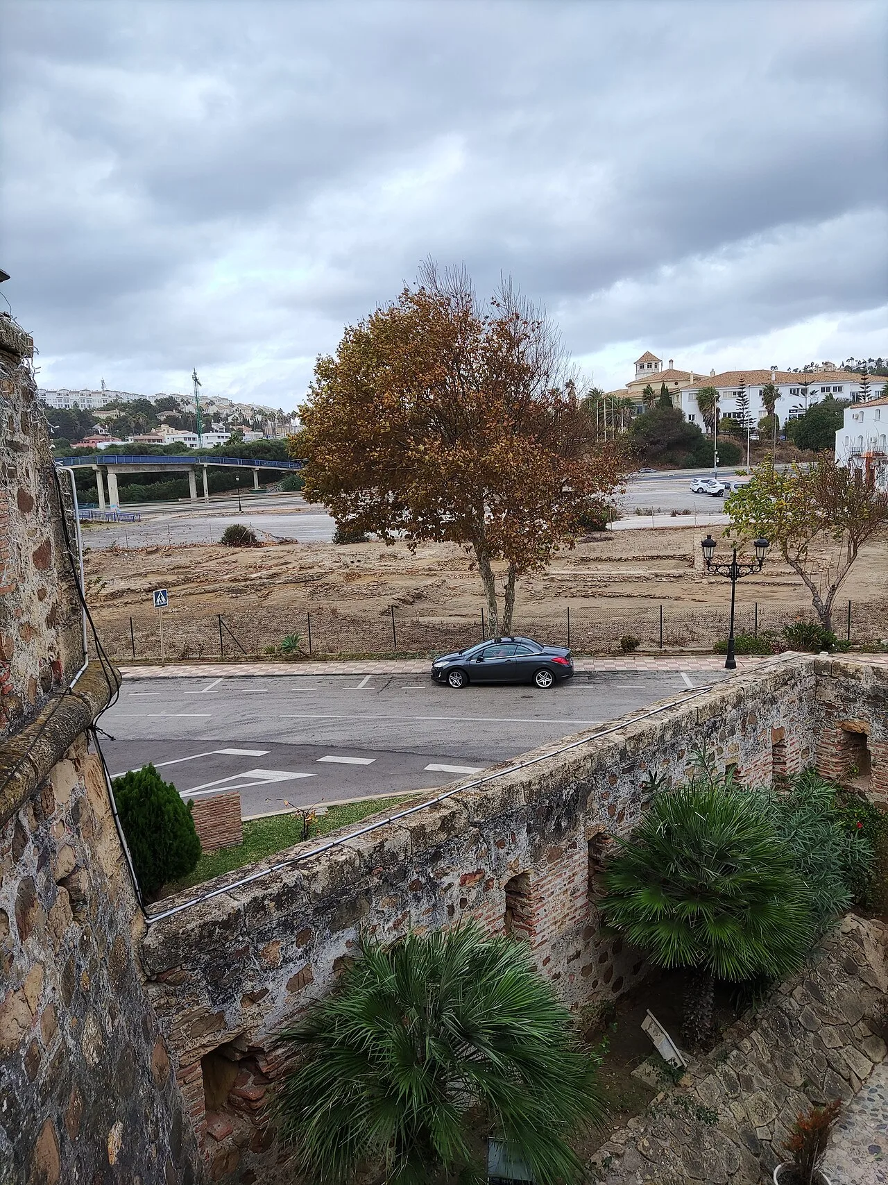 Vista de árboles, un coche y restos de una muralla en Manilva.