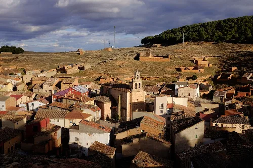 Blick auf Häuser und eine Kirche in Arcos de Jalón mit Hügeln und wolkigem Himmel.