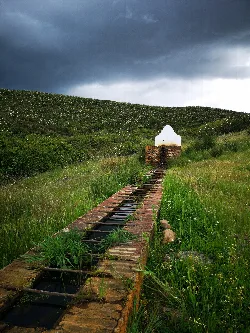 Old fountain with water channel in a green landscape and dark sky.