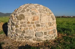 Steinernes Kuppelgebäude in einem Feld mit Bergen und Dorf im Hintergrund.