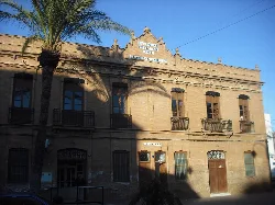 Edificio de dos plantas con ventanas y balcones, con un cartel que indica las escuelas públicas.