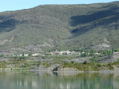 Blick auf das Dorf Arguis am Stausee mit Bergen im Hintergrund.