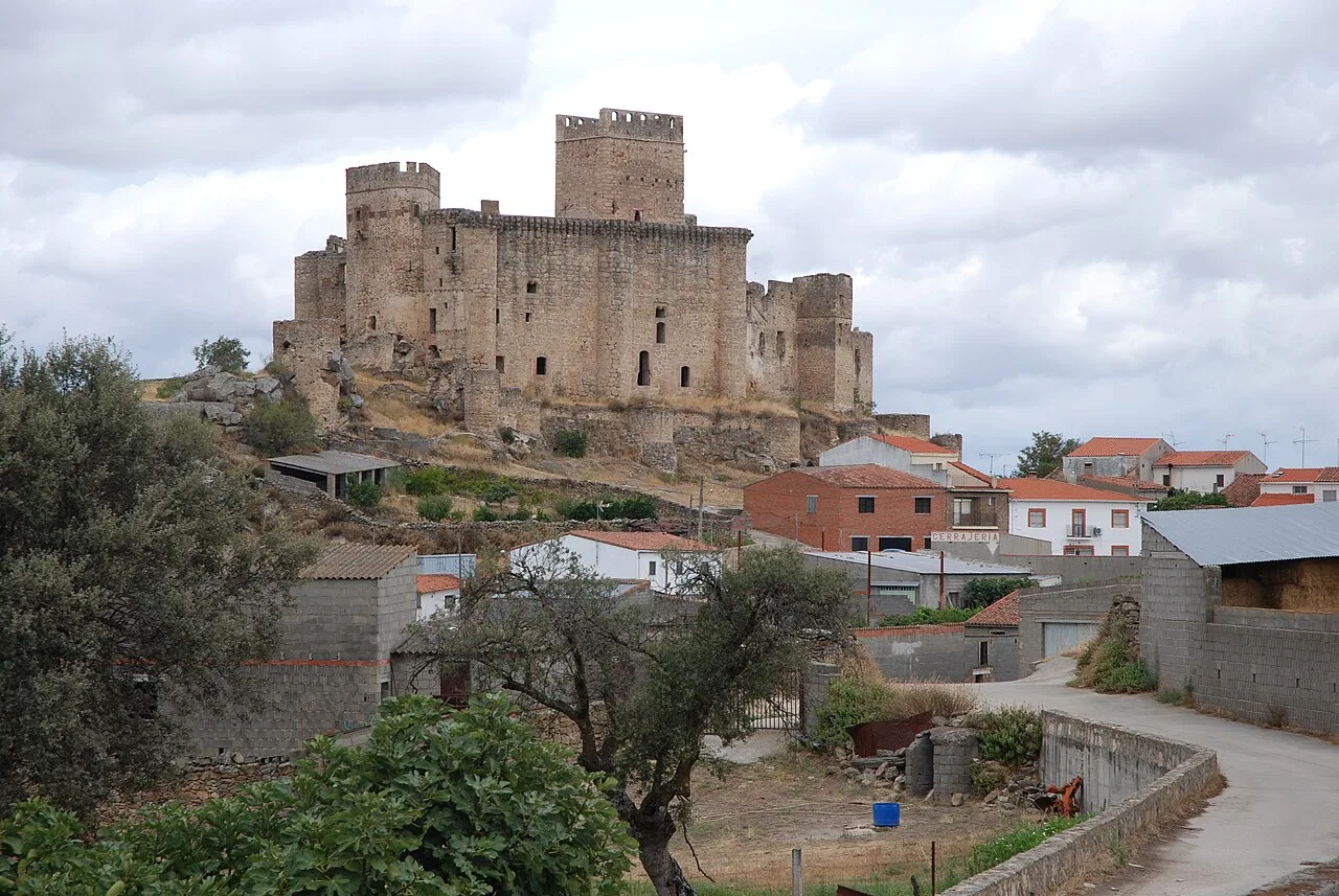 Castillo de piedra con torres en lo alto de una colina, sobre casas y árboles.