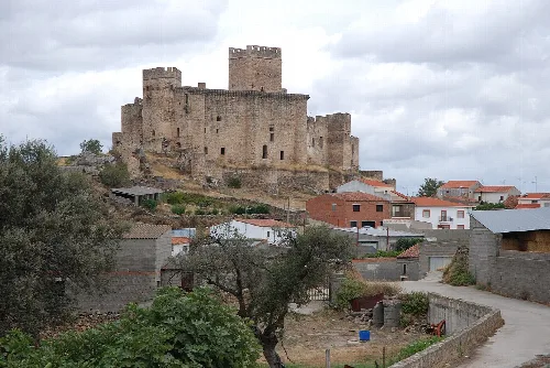 Steinernes Schloss mit Türmen auf einem Hügel über Häusern und Bäumen.
