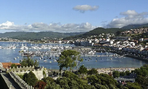 Blick auf den Hafen von Baiona mit Booten und Hügeln im Hintergrund.