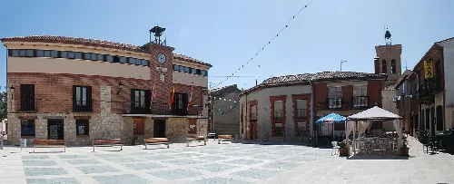 Square in Magaz de Pisuerga with buildings and a terrace with tables and umbrellas.